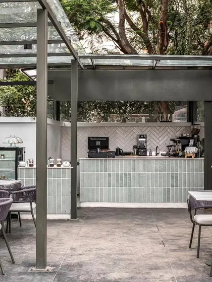 The open-air coffee bar at Kana Cafe in Bangalore, designed to be the heart of the space. The design integrates nature with a breezy, Bali-inspired aesthetic, featuring a glass canopy, minimal furniture, and a dessert counter clad in soft green tiles to create a serene urban retreat.