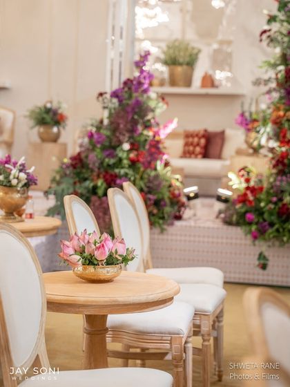 Elegant seating for the "Garden of Vows" ceremony. Classic white chairs with natural wood frames are paired with small tables, each holding a beautiful pink rose arrangement.