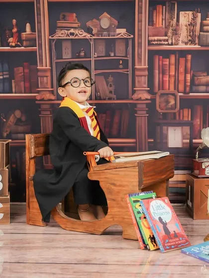 Ready to cast a spell. The vintage school desk and bookshelf backdrop create an authentic Hogwarts classroom feel for this themed baby photoshoot.