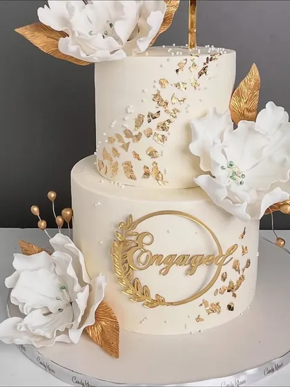 A beautiful two-tiered engagement cake in white and gold, featuring large sugar flowers, gold leaf, and an elegant "Engaged" topper.