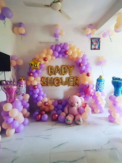 A baby shower decoration in pastel purple and peach, with a balloon ring, foil letters, and a cute teddy bear.