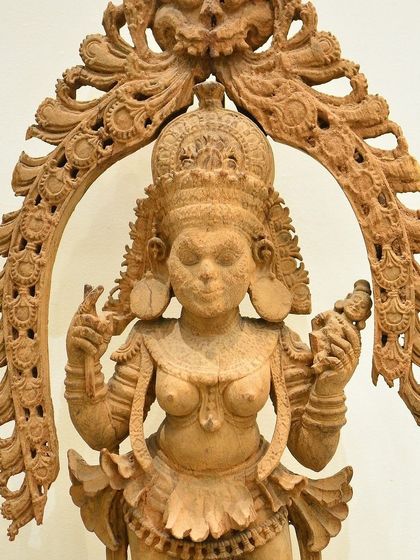 An unpainted wooden sculpture of an Apsara, a celestial dancer. Her poised stance, with the Jnana Mudra in one hand and a mirror in the other, blends wisdom with beauty, reflecting delicate craftsmanship.