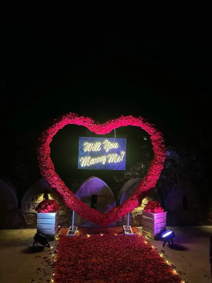 A full view of the stunning red rose heart arch against the night sky. This flower-intensive proposal decoration includes a neon sign, a petal-strewn carpet, and spotlights to highlight the setup.