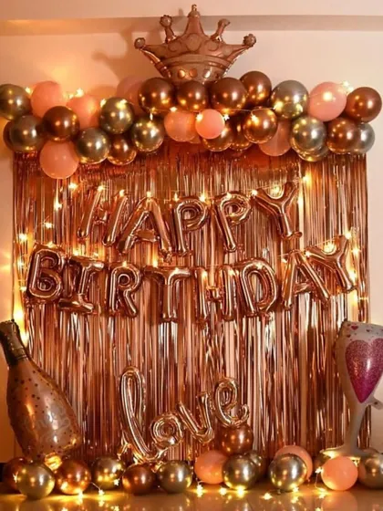 A very popular birthday decoration in a rose gold and gold theme. This setup includes a shiny foil curtain, 'Happy Birthday' letters, a balloon arch topped with a crown, and other fun balloons like a champagne bottle and a 'Love' sign.