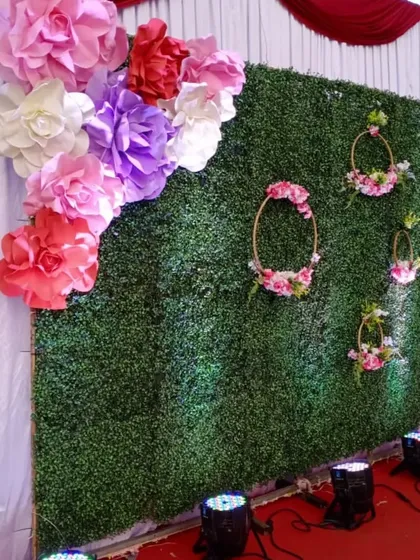 A fresh and vibrant photo wall for a ring ceremony. This backdrop uses an artificial green grass wall as a base, decorated with large, colorful paper flowers and delicate floral hoops for a garden-party feel.