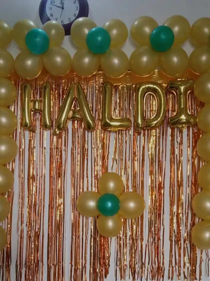 A simple Haldi ceremony backdrop for a home function. The decoration uses flower-shaped balloon patterns and a "Haldi" foil banner against a shimmer curtain.