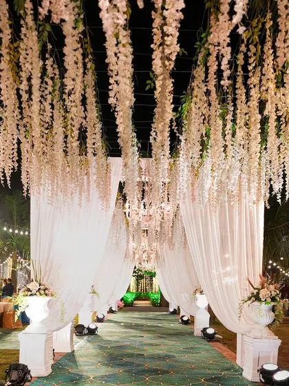A grand entrance for an evening reception. The walkway is lined with white drapes and features a stunning ceiling of hanging white wisteria, creating a dreamy, immersive experience.