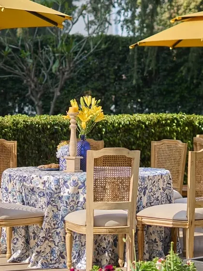 A poolside dining table with cane-back chairs, shaded by a yellow umbrella, and decorated with a simple floral centerpiece.