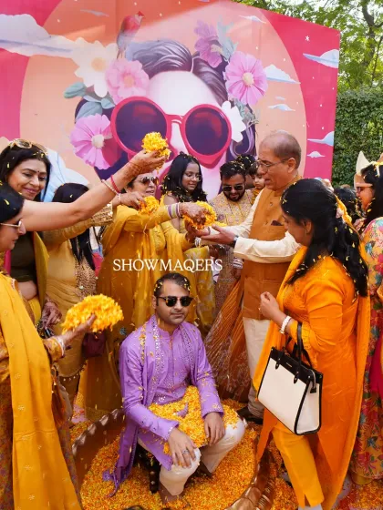 The groom is showered with flower petals by family and friends during the Haldi ceremony, with a colorful and artistic backdrop behind him.