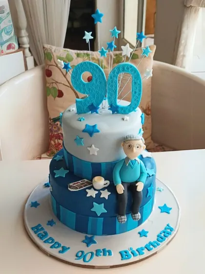 A cheerful two-tier cake for a 90th birthday, decorated in shades of blue with stars and a charming fondant figure of the birthday man.