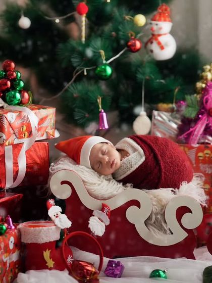 A newborn in a Santa hat sleeps in a miniature sleigh, surrounded by Christmas presents and decorations.