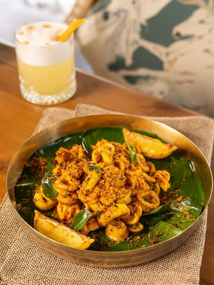 A plate of spicy calamari served on a banana leaf in a brass dish. The styling gives it a coastal, Goan vibe, especially when paired with a refreshing cocktail.