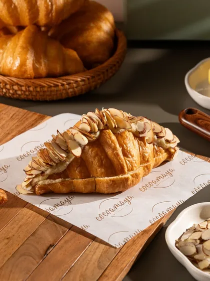 A flaky almond croissant, generously topped with sliced almonds. The lighting catches the golden-brown crust and buttery layers.