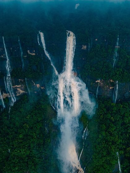 Another stunning aerial view of a massive waterfall in Meghalaya, its white water a stark contrast against the deep green jungle.