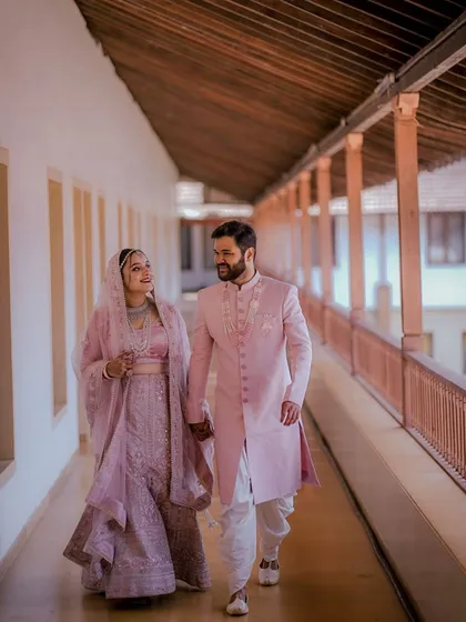 A couple in matching pastel pink outfits walks hand-in-hand down a beautiful, rustic corridor, creating a picture of elegance and romance.