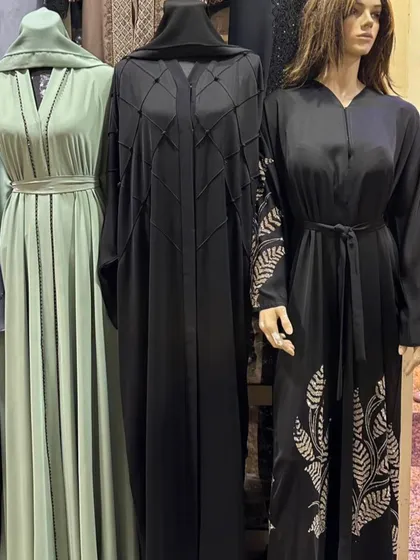 A selection of abayas on mannequins, including a sage green design, a black piece with geometric pintucks, and another with leaf embroidery.