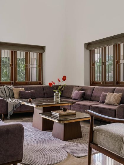 The living room of Relic House is centered around a large, comfortable L-shaped sofa in a muted violet hue. We paired it with solid wood coffee tables and armchairs, creating a warm and inviting space for family gatherings, grounded by a textured rug with a subtle circular pattern.