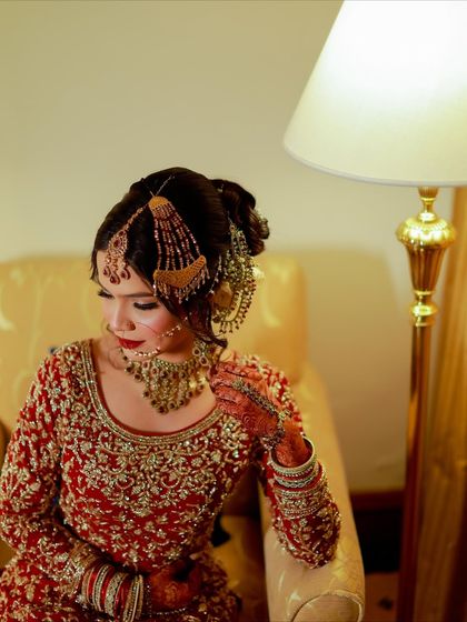 A portrait of a bride with a bold red lip and a soft glam look. Her hair is styled in an elegant updo, adorned with a traditional jhoomar.