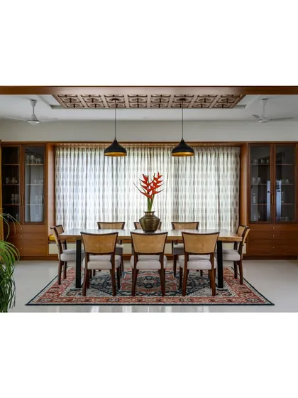 The 'Vasudhalayah' dining area features a live-edge table anchored by an intricately carved wooden ceiling panel. The space is flanked by two crockery units and features a bay window seating nook.