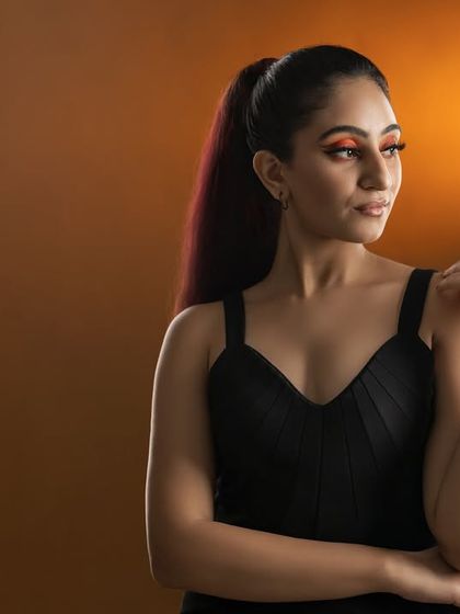 A chic and modern fashion portrait of a model in a black top against a warm studio backdrop, showcasing dramatic makeup and a high-ponytail hairstyle.