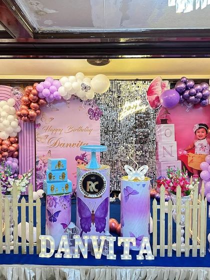 A full view of a butterfly-themed stage decoration with personalized photo cutouts and a purple and metallic balloon arrangement. The setup includes themed plinths and light-up letters for the name.