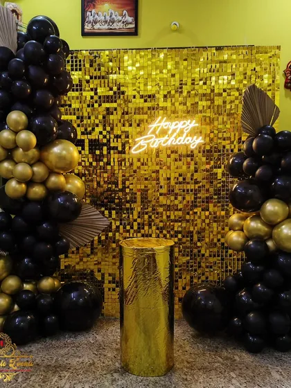 A chic and glamorous birthday setup featuring a dazzling gold shimmer wall as the backdrop. The look is completed with stylish black and gold balloon columns and a modern 'Happy Birthday' neon sign.