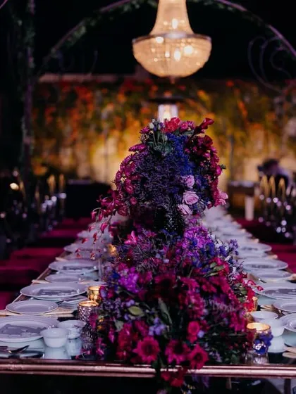 The long table setup for the reception dinner, featuring a lush, dramatic floral runner in deep jewel tones and elegant chandeliers.
