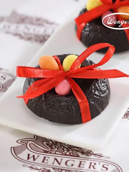 A close-up of our chocolate Easter nest. This delicate creation is made from dark chocolate, shaped like a bird's nest, and filled with tiny, colourful candy eggs, all tied with a red ribbon.