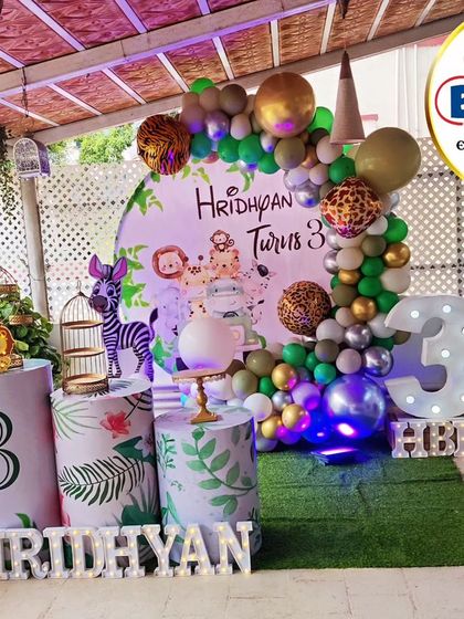 An outdoor jungle-themed setup for a third birthday. This decor features a circular backdrop with animal characters, balloon-animal sculptures, and a faux grass floor, perfect for a garden party.