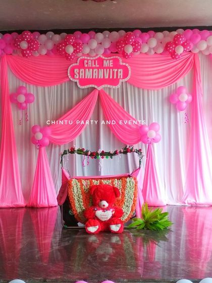 A beautiful naming ceremony stage decoration in pink and white. The setup includes draped curtains, a full balloon arch, and a flower-decorated cradle.