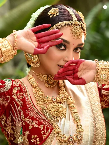 An expressive bridal shot where the model frames her face with her 'alta' stained hands. This pose is both traditional and artistic, drawing attention to her eyes and the intricate gold jewellery.