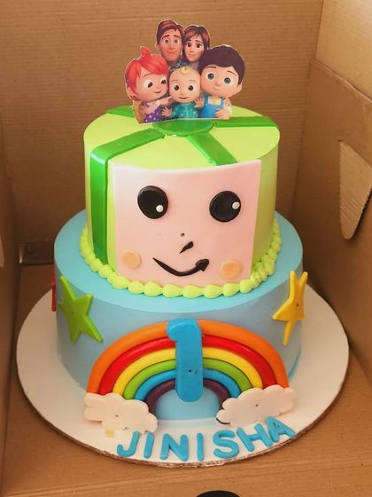 A Cocomelon cake designed to look like a gift box. This two-tier cake is perfect for a first birthday, complete with a rainbow and the Cocomelon family on top.
