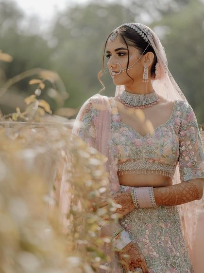 A bride in a pastel floral lehenga stands amidst tall grasses, the soft light creating a gentle and romantic mood.