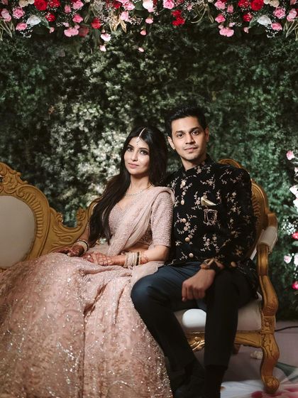 The couple seated against a backdrop of a dense floral wall. The rich greenery provides a beautiful contrast to the soft pinks and whites of the roses, creating a classic and timeless look.