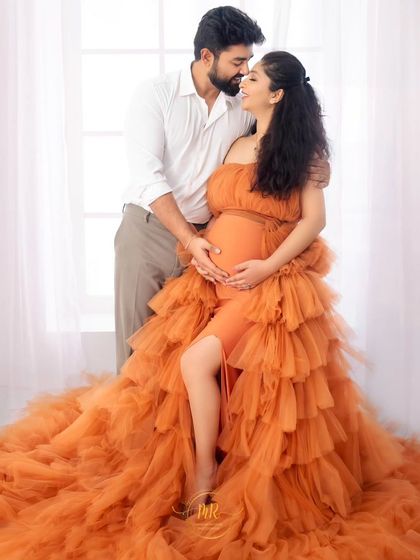 A beautiful full-length portrait of a couple. They share an intimate gaze, with the mother-to-be seated in a stunning orange ruffled gown.