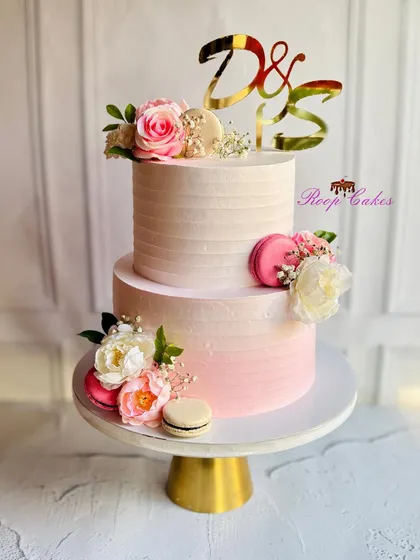 Layers of love in blush, blooms, and gold. This two-tier engagement cake features a soft pink ombre, fresh flowers, and delicate macarons, personalized with the couple's initials on a gold topper.