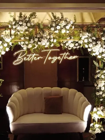 A modern and chic photo booth with a "Better Together" neon sign. The backdrop of white flowers and gold framing is perfect for a stylish engagement or reception party.