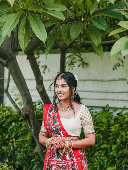 A beautiful portrait of the bride, Aishwarya, in her traditional red and white saree. Her happy, natural smile shines against the lush green backdrop.