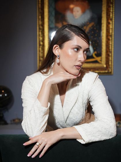A model in a textured white blazer dress, styled for an elegant evening event. The portrait is set against a classic, art-filled interior, adding to the sophisticated mood.