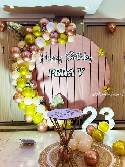 A beautiful and warm-toned birthday setup featuring a striped pink backdrop, an organic balloon garland, and a light-up number '23'.