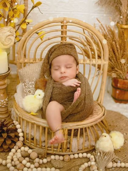 A full view of a rustic, boho-themed newborn setup with the baby sleeping peacefully in a wicker chair, surrounded by dried grasses and pinecones.
