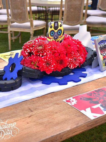 A detailed shot of the table centrepiece, combining bold red flowers with miniature tires, gears, and a framed Transformer image. Every element is carefully chosen to enhance the theme.