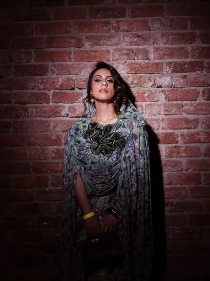 A full-length view of the Anamika Khanna x H&M look. The hair is styled with soft curls, and the makeup is bold enough to stand out against the patterned fabric and dramatic silhouette of the cape.
