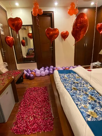 This romantic room setup features a thick pathway of red rose petals leading to the bed. The room is filled with red heart-shaped foil balloons and bunches of regular balloons hanging from the ceiling to create a classic and passionate atmosphere.