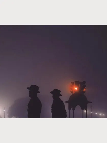 A silhouette of two soldiers against the foggy backdrop of a Delhi winter morning during Republic Day rehearsals. The red traffic light adds a single point of color to the moody, atmospheric shot.