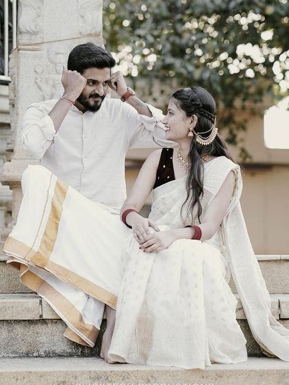 A playful moment between a couple in traditional white South Indian attire, sitting on steps and sharing a laugh.