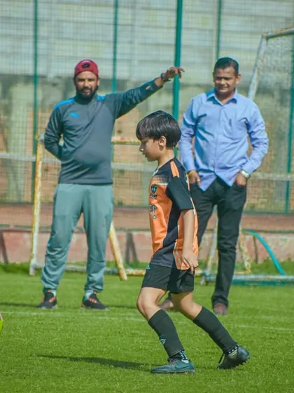 A young player focusing on his technique as he strikes the ball, with a coach guiding from the sidelines.