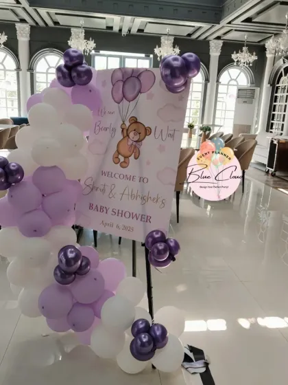A teddy bear themed baby shower welcome sign with a beautiful pastel purple and white balloon garland.