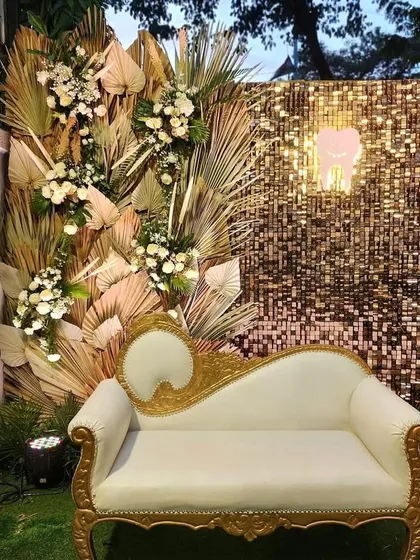 A chic boho photo booth with a gold shimmer wall and dried palm leaf arrangements. The classic white and gold diwan provides an elegant seating option for photos.