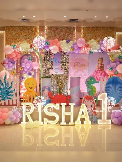 A full view of the enchanting mermaid-themed stage for Risha's first birthday. The setup includes marquee letters, a floral wall, and a variety of sea creature props.